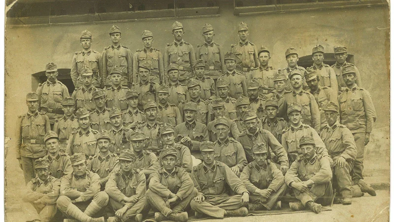 Fotografija: Dopisnica s skupino vojakov v Gleisdorfu pri Gradcu, ki jo je Lovro Kuhar poslal staršem 23. avgusta 1915. Lovro Kuhar stoji točno na sredini, tik nad sedečim stotnikom (verjetno Leo Dettela). Vir: Musilov inštitut v Celovcu