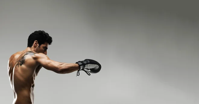 Tattooed muscular male fighter punching in the air against plain background