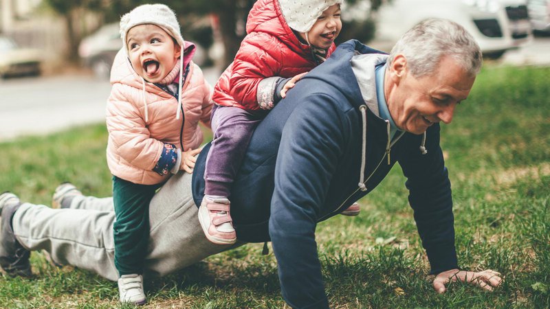 Fotografija: A photo of a playful grandfather and granddaughter. They are casually dressed and playing in the park. They exercise together. A grandfather is exercising while granddaughters are sitting on his back.
