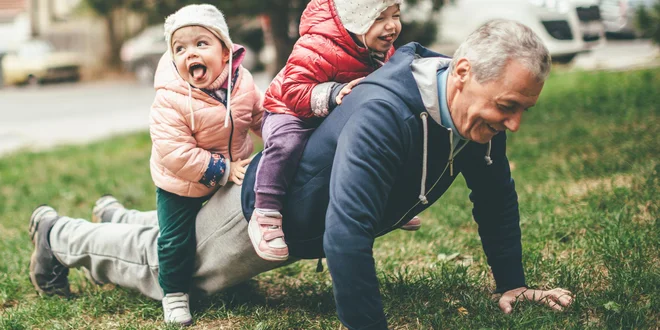 A photo of a playful grandfather and granddaughter. They are casually dressed and playing in the park. They exercise together. A grandfather is exercising while granddaughters are sitting on his back.
