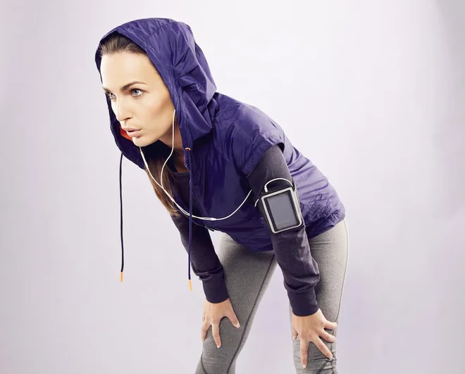 Young caucasian woman taking breath after jogging. Female athlete resting with hands on knees and looking away over grey background