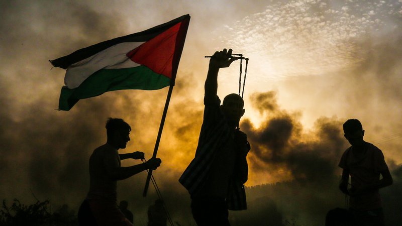 Fotografija: Palestinski demonstrant drži nacionalno zastavo, ko drugi protestnik meče kamen proti izraelskim silam med spopadi na plaži blizu morske meje z Izraelom na severu Gaze. Foto Mahmud Hams Afp
