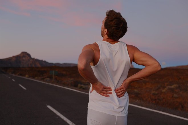 Back pain. Athletic running man with injury in sportswear rubbing touching lower back muscles standing on road outside at night.