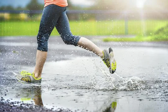 Young woman running on asphalt sports field in rainy weather. Details of legs and sports shoes splashing in puddles.
