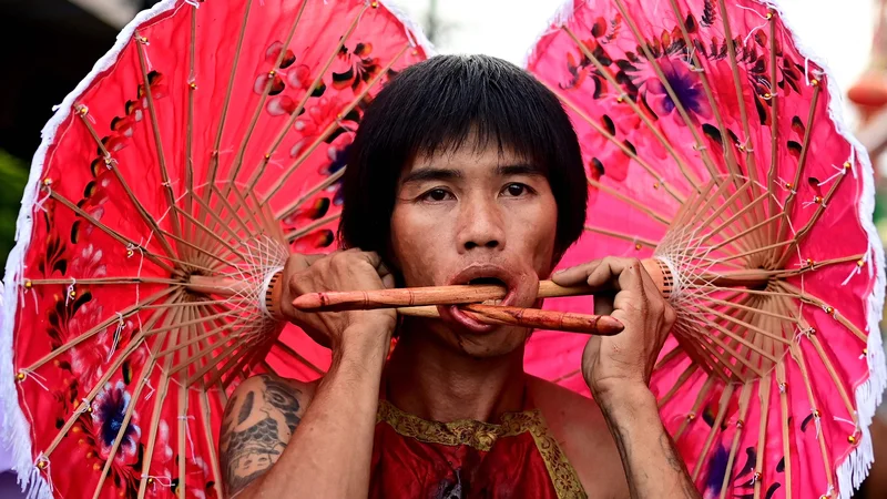 Fotografija: Tajski častilec svetišča Kuan Tae Kun s papirnatimi dežniki, prebodenimi skozi lica, sodeluje v procesiji med letnim vegetarijanskim festivalom v Phuketu. Foto: Manan Vatsyayana/Afp
