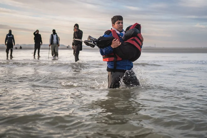 Migrant nosi otroka, za njim pa sobegunci, ki se želijo vkrcati na čoln na plaži Gravelines blizu Dunkerqueja, na severu Francije, ko poskušajo prečkati Rokavski zaliv. Od začetka leta je nevarno prečkanje Rokavskega preliva, ene od najbolj prometnih ladijskih poti na svetu, po kateri vsak dan pluje več kot 400 komercialnih ladij, opravilo že več kot 33500 ljudi. Foto: Sameer Al-doumy/Afp