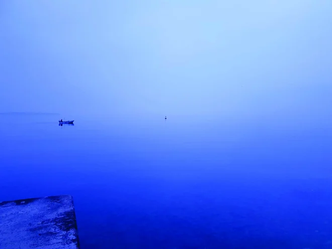 Megla s svojo neskončnostjo, kjer se horizont zamegli in ni več jasne ločnice med ravnino morja in širino neba, bolj ali manj vse postane sivina ali modrina, ki pa jo kot detajl osredotoča ribiški čoln.

V Piranu pozimi 2022.

FOTO (in opis iz knjige): Robert Potokar
