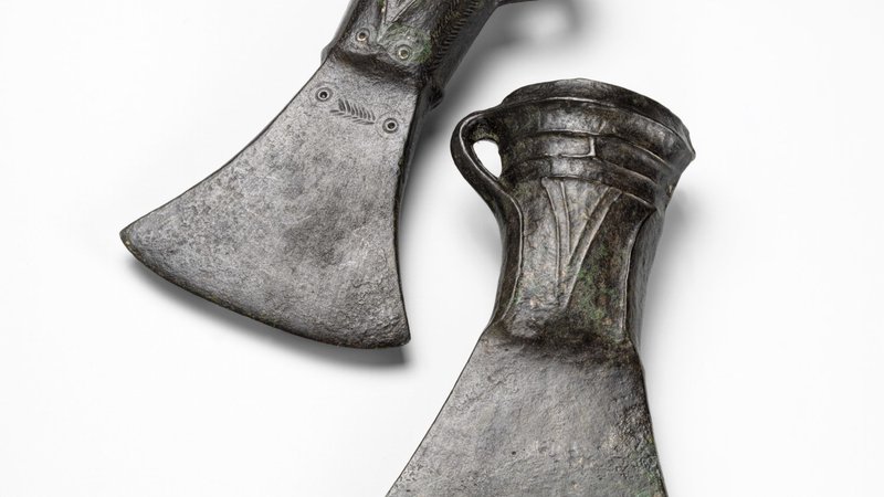 Fotografija: V pozni bronasti dobi so se v srednjo Evropo z vzhoda razširile bronaste tulaste sekire. Pri teh sekirah je bilo nasadišče vdeto v cevast tul in dodatno pričvrščeno z jermenjem, privezanim na stransko ušesce. Sekiri iz Ljubljanice pri Bevkah iz 11.–10. stoletja pr. n. št. sta v času kulture žarnih grobišč mlajši različici tulastih sekir, bogato okrašeni z ulitim rebrastim in vtolčenim okrasom. FOTO: Tomaž Lauko ©Narodni muzej Slovenije