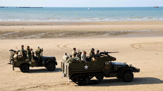 Navdušenci, oblečeni v replike vojaških oblačil iz obdobja druge svetovne vojne, se vozijo na džipu Willys in ameriškem poltovornjaku M3 na plaži v Arromanches-les-Bains v severozahodni Franciji v okviru spominskih slovesnosti ob 80. obletnici izkrcanja zaveznikov v Normandiji v času druge svetovne vojne. Foto: Ludovic Marin/Afp