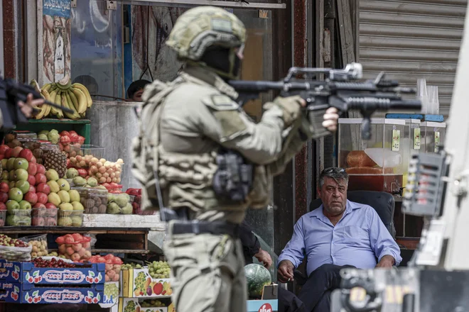 Vojak izraelske vojske meri s puško, medtem ko ga Palestinec opazuje med racijo v središču palestinskega mesta Nablus na okupiranem Zahodnem bregu. Izraelske sile so v torek v več mestih na Zahodnem bregu, vključno z Ramalo in Nablusom, izvedle racijo v menjalnicah, pri čemer so njihovo matično podjetje obtožile povezav s terorističnimi organizacijami. Foto: Jaafar Ashtiyeh/Afp