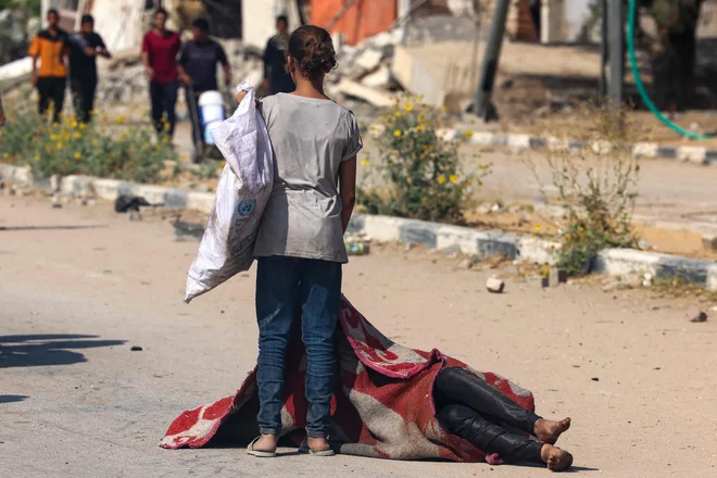 Palestinska deklica stoji nad pokritim truplom osebe, ki je bila ubita med čakanjem na hrano v razdelilnem mestu, ki ga upravlja skupina Gaza Humanitarian Foundation (GHF), v Nusseiratu v osrednjem delu Gaze. Humanitarne organizacije so opozorile, da prebivalstvo Gaze čaka katastrofalna lakota, ki jo je sprožila izraelska omejitev pomoči. Foto: Afp